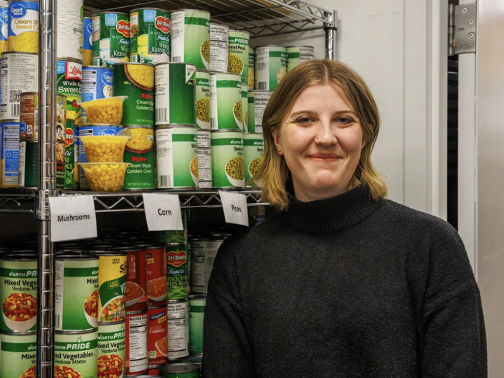 Young woman, Kaitlyn Richards, standing in front of a shelf with canned goods. She has short blonde hair and is smiling while wearing a black sweater.